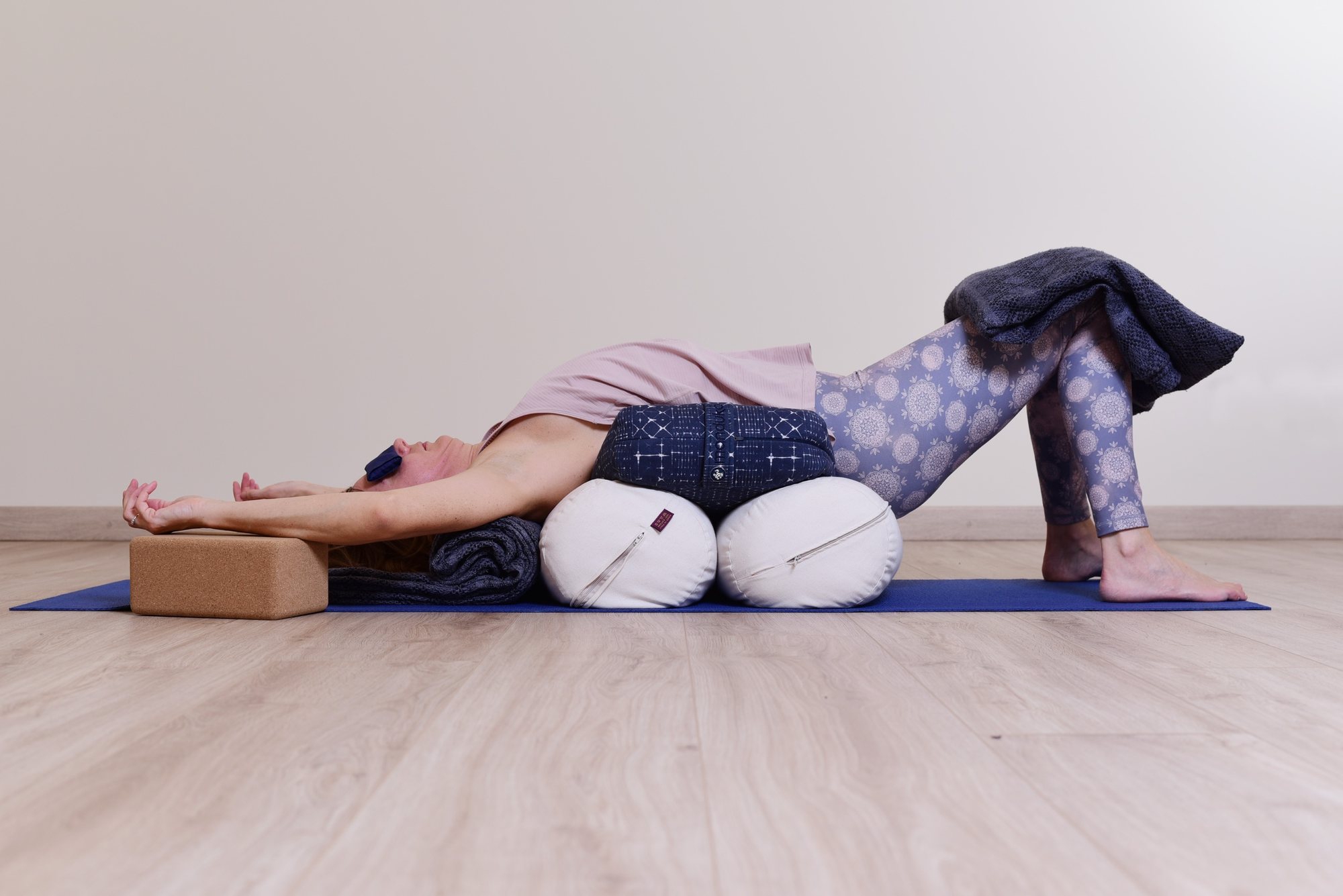 Flessione all’indietro sostenuta: la schiena su due bolster rotondi e un cuscino, le braccia distese sopra la testa su un mattoncino, le ginocchia piegate sotto una coperta e un cuscino sugli occhi